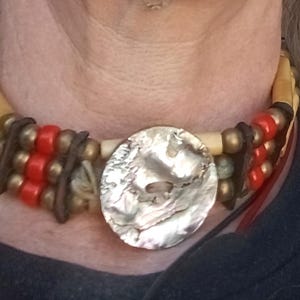 May include: A choker necklace with multiple strands, incorporating bone-coloured beads, red beads, and a large, round, iridescent button. The necklace has a tribal or bohemian style, with leather accents and a natural colour scheme.