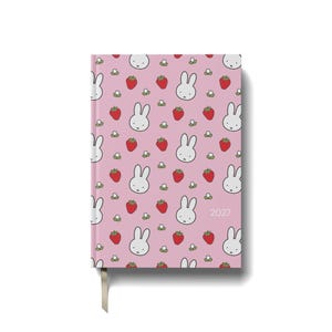 May include: A pink hardback planner featuring a repeating pattern of white rabbit faces, red strawberries, and small white flowers. The year 2027 is printed in small white text on the front cover, which includes a fabric ribbon bookmark.