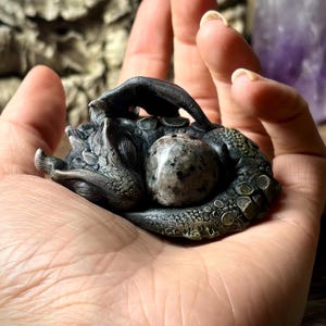 May include: A small, detailed, silver-colored dragon sculpture curled around a grey and white speckled stone. The sculpture is resting in the palm of a hand.
