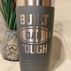 May include: A dark gray stainless steel tumbler with a silver lid. The tumbler has the words "BUILT DAD TOUGH" engraved on it.