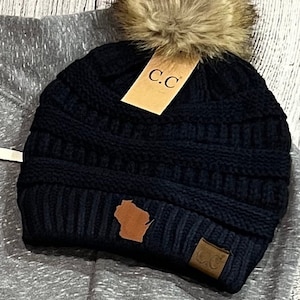 May include: Black knit beanie with a brown faux fur pom pom, a brown leather patch with the state of Wisconsin, and a brown leather patch with the letters "C.C."