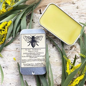 May include: A small tin container with a silver lid and a label that reads "The Hive - Handmade on Magnetic Island Sandalwood Vanilla Solid Perfume". The label also includes instructions for use and a list of ingredients. The tin is open and a yellow balm is visible inside. The tin is sitting on a wooden surface with green leaves and yellow flowers.