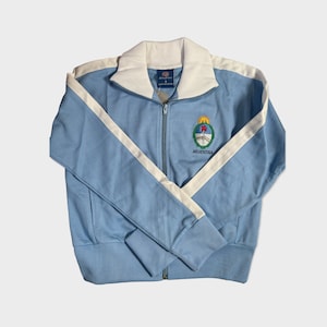 May include: A light blue zip-up jacket with white stripes down the sleeves and a white collar. The jacket has an embroidered crest with the word "ARGENTINA" underneath.