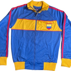Op de afbeelding: Een blauw en geel trainingsjack met rode piping. Het jack heeft een ritssluiting en een gele en rode streep over de borst. Het woord "COLOMBIA" staat op de borst gedrukt met een geel en rood schild.