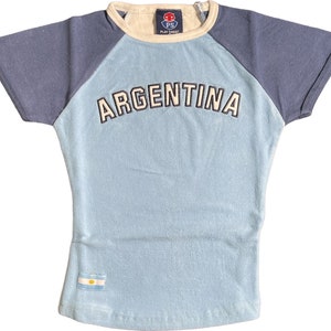 Peut inclure: Un t-shirt bleu clair avec un design de manches raglan bleu marine. Le t-shirt a un col rond blanc et le mot "ARGENTINA" en lettres blanches sur le devant. Un petit drapeau bleu et blanc se trouve en bas du t-shirt.