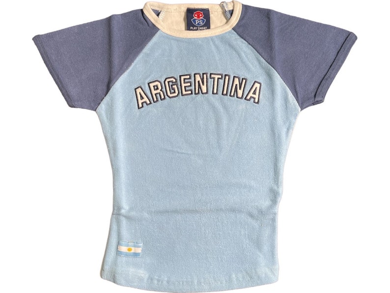Peut inclure: Un t-shirt bleu clair avec un design de manches raglan bleu marine. Le t-shirt a un col rond blanc et le mot "ARGENTINA" en lettres blanches sur le devant. Un petit drapeau bleu et blanc se trouve en bas du t-shirt.