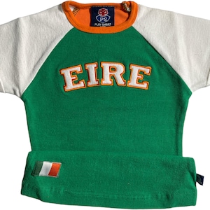 Può includere: Una maglietta a maniche corte verde e bianca con un bordo arancione e la parola "EIRE" ricamata sul davanti. La maglietta ha una toppa della bandiera irlandese bianca e verde sul lato inferiore sinistro.