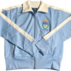 May include: A light blue zippered jacket with white stripes down the sleeves and a white collar. The jacket has an embroidered crest with the word "ARGENTINA" underneath.