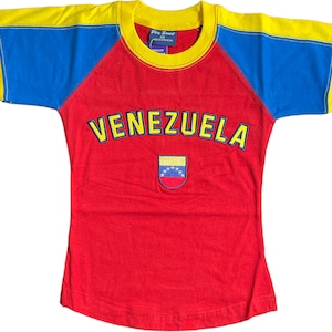 May include: A red, yellow, and blue t-shirt with a baseball style design. The shirt has the word "VENEZUELA" in yellow letters on the front and a small embroidered Venezuelan flag.