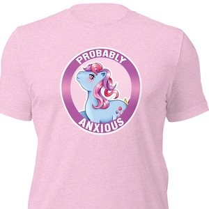 May include: Pale pink t-shirt with a cartoon pony illustration. The pony is blue with pink hair, inside a purple circle with the words "PROBABLY ANXIOUS". The shirt has a crew neck and short sleeves.