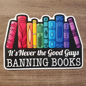 May include: A sticker featuring a row of colorful books with the text "It's Never the Good Guys BANNING BOOKS" in white on a black background. The books are in various colors, including pink, red, orange, yellow, green, blue, and purple.