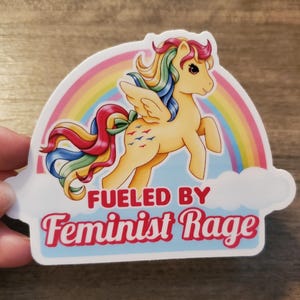 May include: A die-cut sticker featuring a light yellow winged pony with a multi-colored rainbow mane and tail. Behind the pony is a colorful rainbow and a white cloud. The text FUELED BY Feminist Rage is printed in red below the pony.