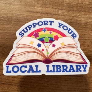 Puede incluir: Una pegatina con el texto "SUPPORT YOUR LOCAL LIBRARY" en azul, arqueado sobre un libro abierto con un arcoíris y estrellas. El libro tiene páginas color beige y lomo morado, sobre fondo de madera.
