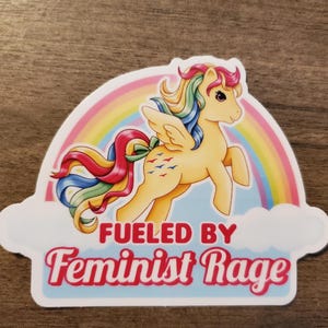 May include: A die-cut sticker featuring a yellow winged pony with a multi-colored rainbow mane and tail, positioned in front of a large rainbow and white clouds. The text FUELED BY Feminist Rage is printed in bold red and pink lettering below.