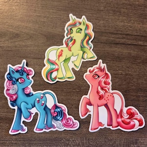 Könnte beinhalten: Drei bunte Einhorn-Aufkleber. Einer ist hellgrün mit Regenbogenhaar und einem roten Vogel. Ein anderer ist blau mit rosa und blauem Haar. Der letzte ist rosa mit rotem Haar und Sternen.