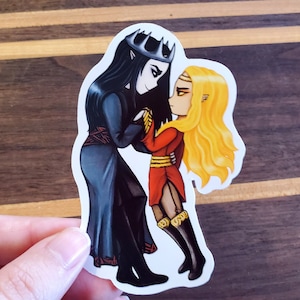May include: A sticker featuring a black-haired character with a crown and a blonde-haired character with a crown. The characters are holding hands and looking at each other. The sticker is on a wooden surface.