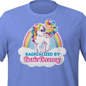 May include: A light blue t-shirt featuring a cartoon pony with a rainbow mane and tail, standing in front of a rainbow. The text "RADICALIZED BY Basic Decency" is printed below the pony in white and red.