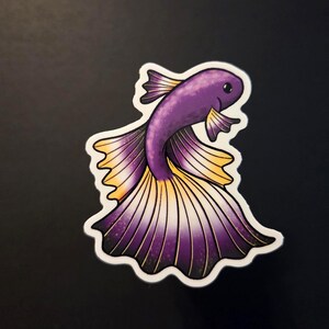 Op de afbeelding: Een sticker met een gestileerde paarse en gele betta-vis met een witte rand. De vis heeft gedetailleerde vinnen en een lachend gezicht. De sticker staat op een zwarte achtergrond.