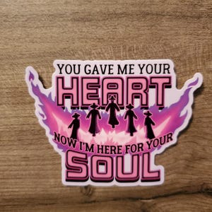 May include: A sticker featuring the text "YOU GAVE ME YOUR HEART, NOW I'M HERE FOR YOUR SOUL" in pink and white. The design includes black cloaked figures and a pink and purple flame-like background.