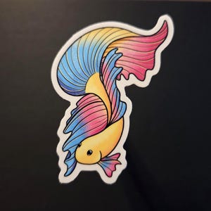 Op de afbeelding: Een kleurrijke sticker met een betta-vis met een vloeiende staart. De vis heeft een gele body en een staart met blauwe, roze en gele secties. De sticker heeft een witte rand en staat tegen een zwarte achtergrond.