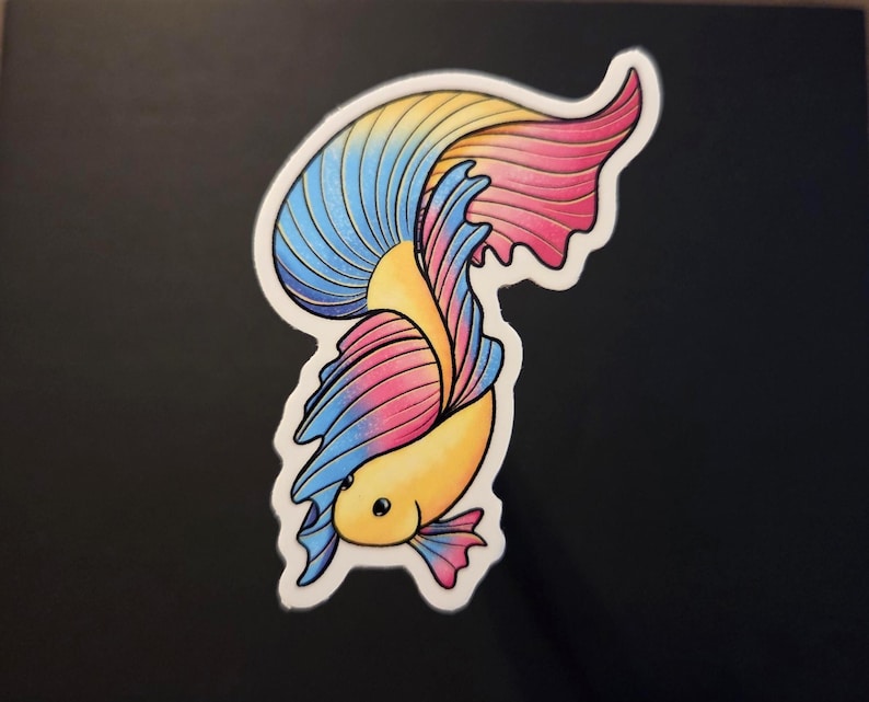 Op de afbeelding: Een kleurrijke sticker met een betta-vis met een vloeiende staart. De vis heeft een gele body en een staart met blauwe, roze en gele secties. De sticker heeft een witte rand en staat tegen een zwarte achtergrond.