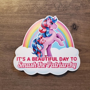 Può includere: Un adesivo con un pony rosa dei cartoni animati con capelli blu e rossi, in piedi davanti a un arcobaleno. Il testo "IT'S A BEAUTIFUL DAY TO Smash the Patriarchy" è scritto su una nuvola bianca.