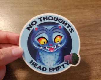 May include: A circular sticker with a blue cat illustration, yellow eyes, and its tongue out. The text reads "NO THOUGHTS" and "HEAD EMPTY" around the cat. A small potted cactus is also depicted on the sticker.
