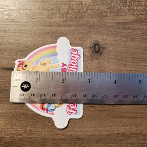 May include: A die-cut sticker featuring a yellow pony with a pink mane, a rainbow, and the text "Feelin' Baby Rage" in pink. A metal ruler is placed across the sticker, showing it measures approximately 3.5 inches in width at its widest point.