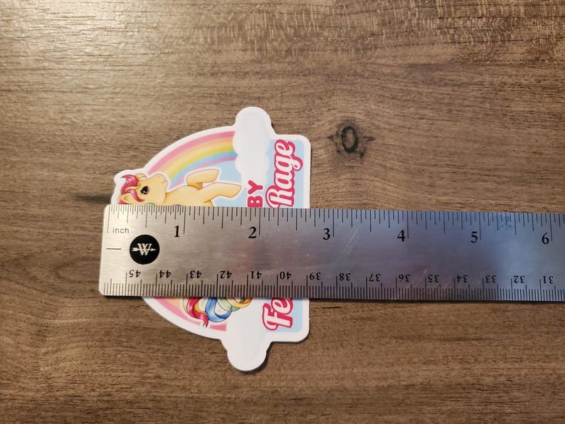 May include: A die-cut sticker featuring a yellow pony with a pink mane, a rainbow, and the text "Feelin' Baby Rage" in pink. A metal ruler is placed across the sticker, showing it measures approximately 3.5 inches in width at its widest point.