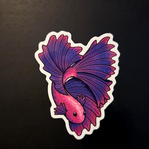 Op de afbeelding: Een sticker met een gestileerde betta-vis in hartvorm. De vis heeft een roze lichaam en grote, vloeiende vinnen in tinten paars en blauw. De sticker heeft een witte rand en staat tegen een donkere achtergrond.