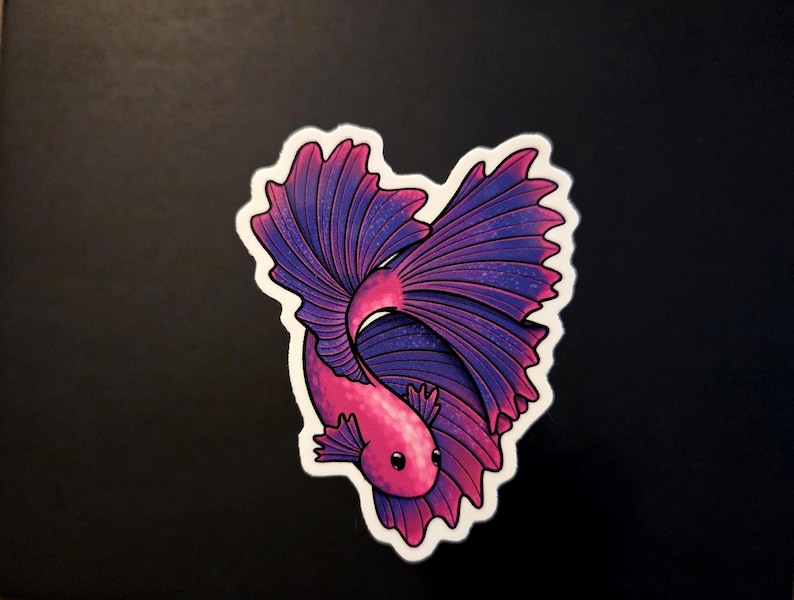 Op de afbeelding: Een sticker met een gestileerde betta-vis in hartvorm. De vis heeft een roze lichaam en grote, vloeiende vinnen in tinten paars en blauw. De sticker heeft een witte rand en staat tegen een donkere achtergrond.