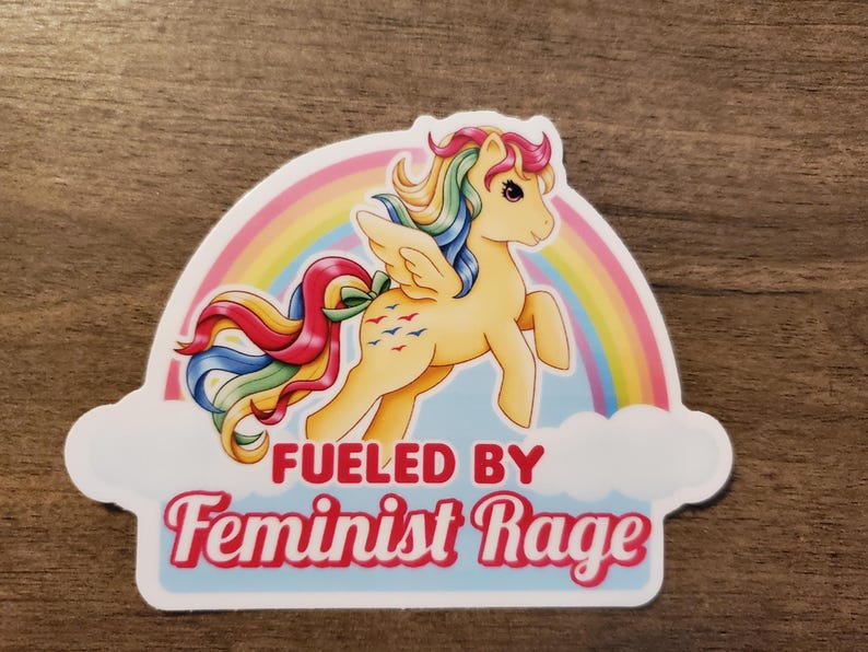 May include: A die-cut sticker featuring a yellow winged pony with a multi-colored rainbow mane and tail, positioned in front of a large rainbow and white clouds. The text FUELED BY Feminist Rage is printed in bold red and pink lettering below.