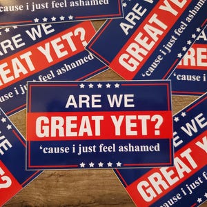 Op de afbeelding: Verschillende rechthoekige stickers met een blauw en rood ontwerp. De tekst "ARE WE GREAT YET?" staat in wit en rood, met de zin "cause I just feel ashamed" eronder.