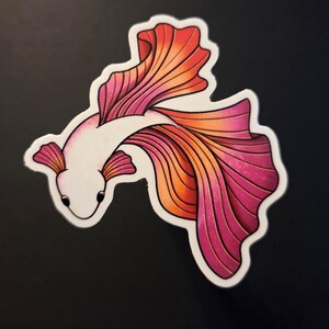 Op de afbeelding: Een sticker met een gestileerde betta-vis met een wit lichaam en vloeiende vinnen in tinten roze, oranje en rood. De vis is omlijnd in zwart en heeft kleine, donkere ogen. Op een zwarte achtergrond.