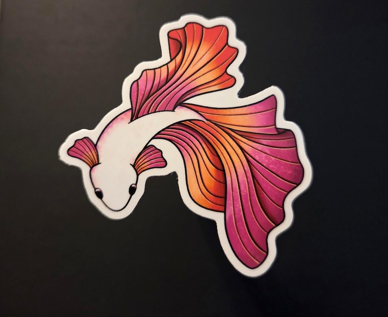 Op de afbeelding: Een sticker met een gestileerde betta-vis met een wit lichaam en vloeiende vinnen in tinten roze, oranje en rood. De vis is omlijnd in zwart en heeft kleine, donkere ogen. Op een zwarte achtergrond.