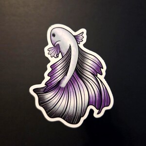 Op de afbeelding: Een sticker met een gestileerde betta-vis met een vloeiende staart. De vis heeft een wit lichaam, een paarse en grijze staart en een witte omtrek. De sticker ligt op een zwart oppervlak.
