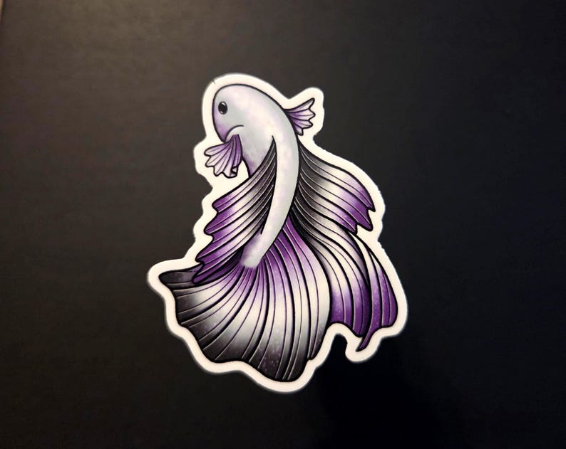 Op de afbeelding: Een sticker met een gestileerde betta-vis met een vloeiende staart. De vis heeft een wit lichaam, een paarse en grijze staart en een witte omtrek. De sticker ligt op een zwart oppervlak.