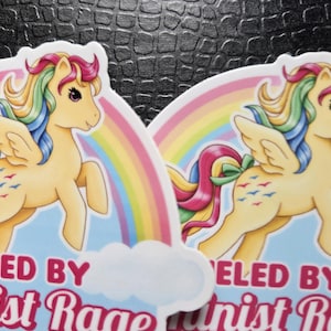May include: Two die-cut stickers featuring a cream-colored winged pony with a rainbow mane and tail. The pony has a green bow on its tail and blue bird symbols on its flank. Behind the pony is a rainbow and the text FUELED BY Feminist Rage.
