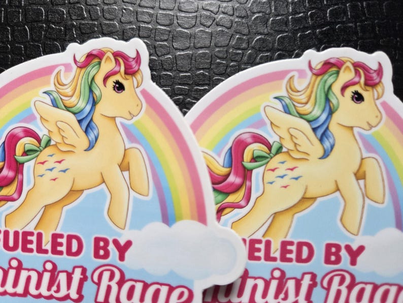 May include: Two die-cut stickers featuring a cream-colored winged pony with a rainbow mane and tail. The pony has a green bow on its tail and blue bird symbols on its flank. Behind the pony is a rainbow and the text FUELED BY Feminist Rage.
