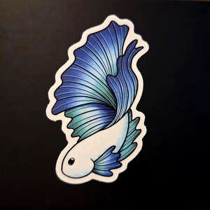 Op de afbeelding: Een sticker met een gestileerde betta-vis, met een wit lichaam en een vloeiende staart. De staart loopt over van blauw naar turquoise, met gedetailleerde lijnen. De sticker heeft een witte rand en staat op een zwarte achtergrond.