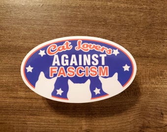 Puede incluir: Pegatina ovalada con fondo azul y el texto "Cat Lovers AGAINST FASCISM" en blanco y rojo. Presenta tres siluetas de gatos blancos y estrellas.