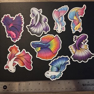 Op de afbeelding: Een verzameling van negen kleurrijke betta vis stickers op een zwart oppervlak. De stickers hebben verschillende ontwerpen met levendige tinten roze, paars, blauw, geel en oranje. Een liniaal is onderaan de afbeelding geplaatst.