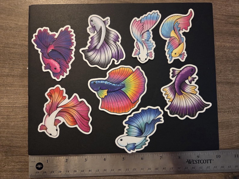 Op de afbeelding: Een verzameling van negen kleurrijke betta vis stickers op een zwart oppervlak. De stickers hebben verschillende ontwerpen met levendige tinten roze, paars, blauw, geel en oranje. Een liniaal is onderaan de afbeelding geplaatst.