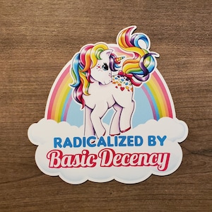 May include: A sticker depicting a white unicorn with a rainbow mane and tail, set against a rainbow. The text "RADICALIZED BY Basic Decency" is written on a cloud-shaped background.
