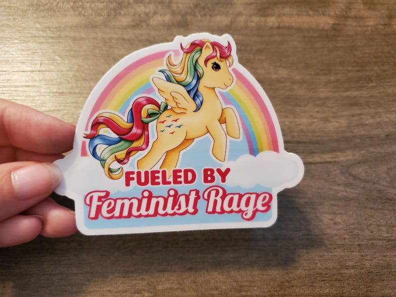 May include: A die-cut sticker featuring a light yellow winged pony with a multi-colored rainbow mane and tail. Behind the pony is a colorful rainbow and a white cloud. The text FUELED BY Feminist Rage is printed in red below the pony.
