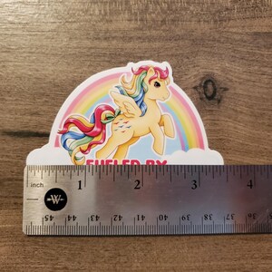 May include: A sticker featuring a yellow winged pony with a rainbow mane and tail, positioned in front of a rainbow and clouds. The text "FUELED BY" is visible at the bottom. A metal ruler shows the sticker is approximately 3.5 inches wide.