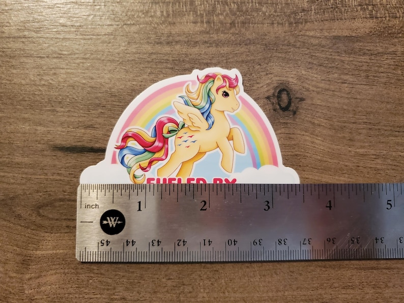 May include: A sticker featuring a yellow winged pony with a rainbow mane and tail, positioned in front of a rainbow and clouds. The text "FUELED BY" is visible at the bottom. A metal ruler shows the sticker is approximately 3.5 inches wide.