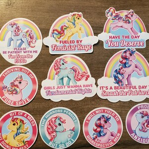 May include: A collection of ten circular and shaped vinyl stickers featuring colorful cartoon ponies with rainbow manes and tails. Each sticker includes text such as "Fueled by Feminist Rage," "Have the Day You Deserve," and "I'm From the 1900s," set against rainbow backgrounds.