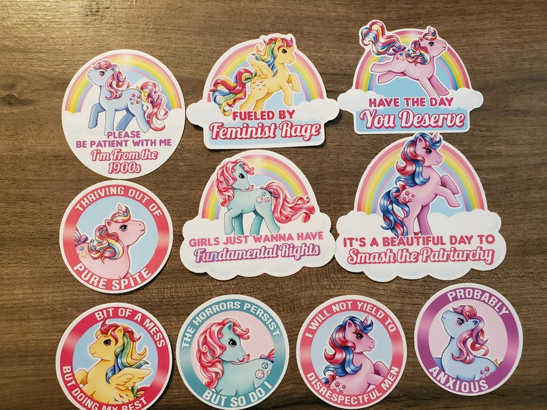 May include: A collection of ten circular and shaped vinyl stickers featuring colorful cartoon ponies with rainbow manes and tails. Each sticker includes text such as "Fueled by Feminist Rage," "Have the Day You Deserve," and "I'm From the 1900s," set against rainbow backgrounds.