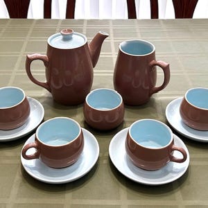 May include: A brown ceramic tea set with a teapot, two mugs, and six cups and saucers. The interior of the cups and mugs is light blue. The set is arranged on a green tablecloth.